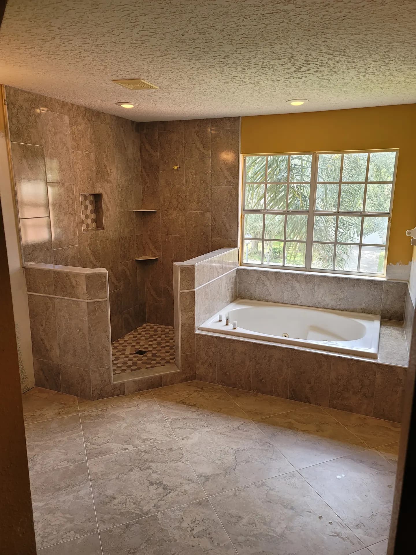 Remodeled master bathroom with large-format porcelain tile on the floor and walls, a tiled tub deck under a bright window, and a walk-in shower with a hexagon mosaic floor.