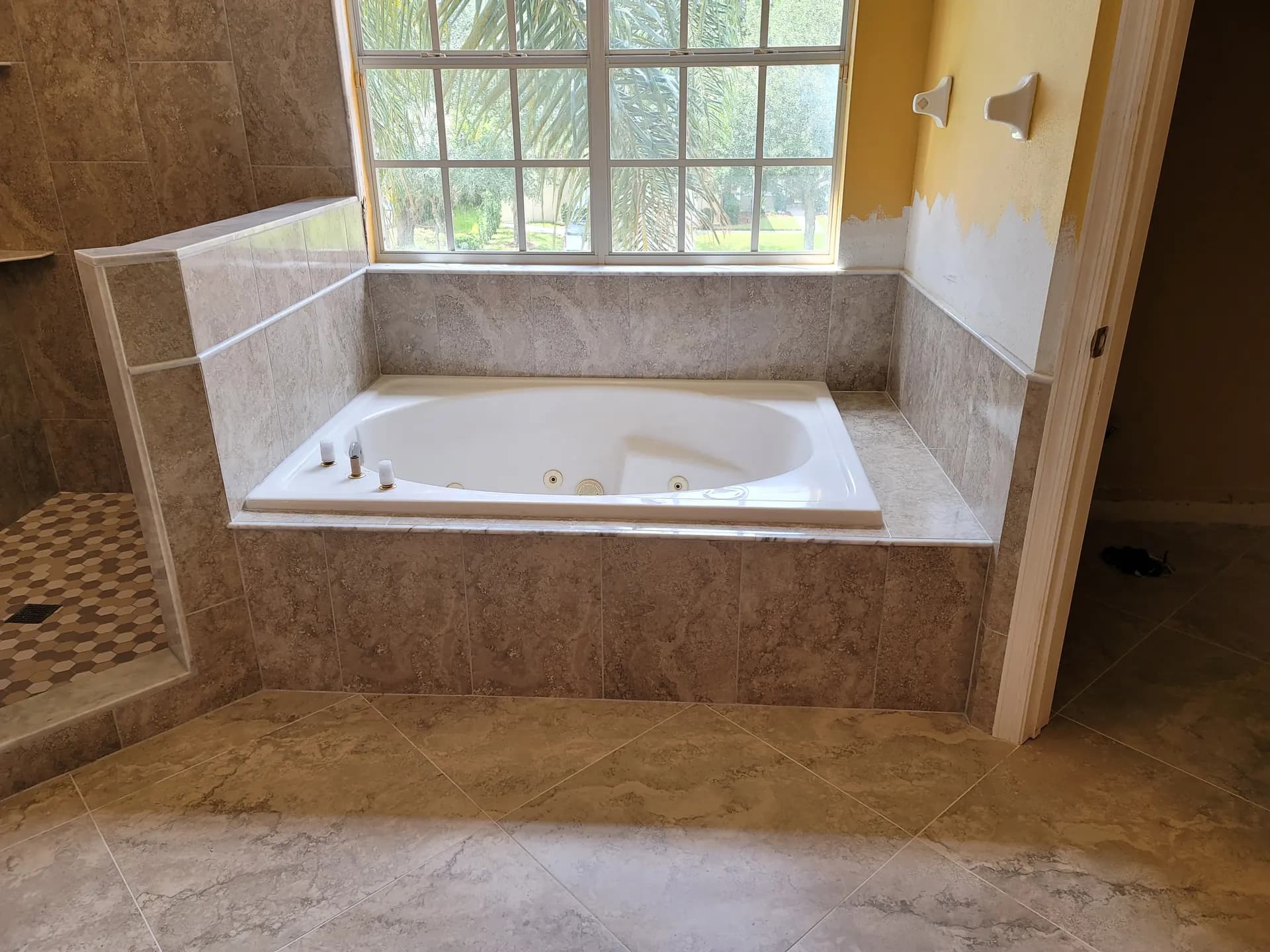 Whirlpool garden tub framed by a marble-look porcelain tile surround and ledge beneath a large window, with diagonally laid large-format floor tile and a hexagon mosaic shower floor visible at left.