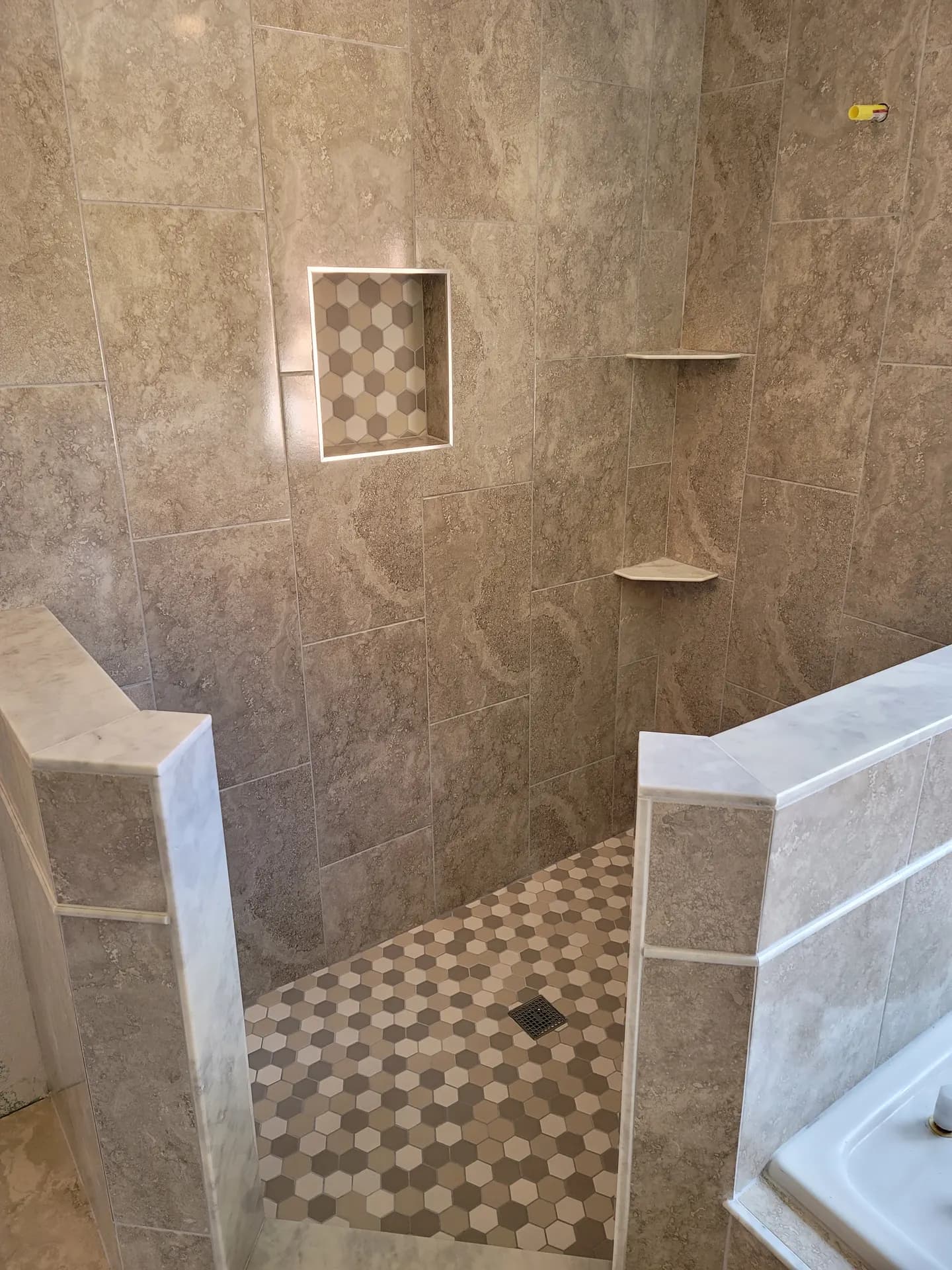 Detail of a walk-in shower with a recessed niche lined in hexagon mosaic tile, corner shelves, marble curb caps, and a matching hex mosaic shower floor.