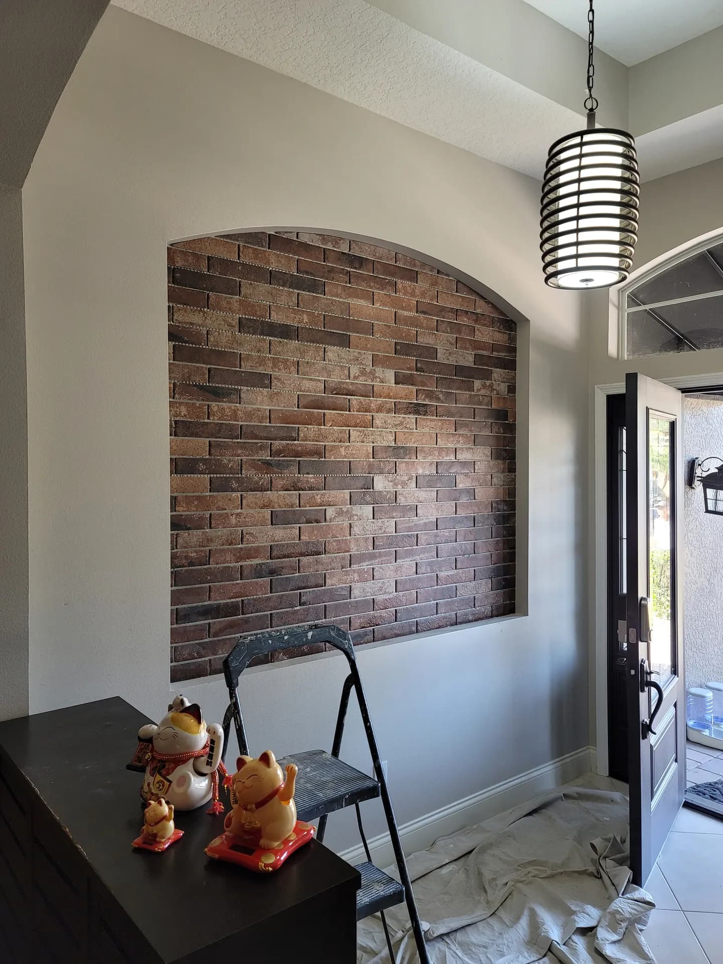 Arched entryway accent wall finished in rustic brick-look mosaic tile beside a black front door, with a caged pendant light overhead.