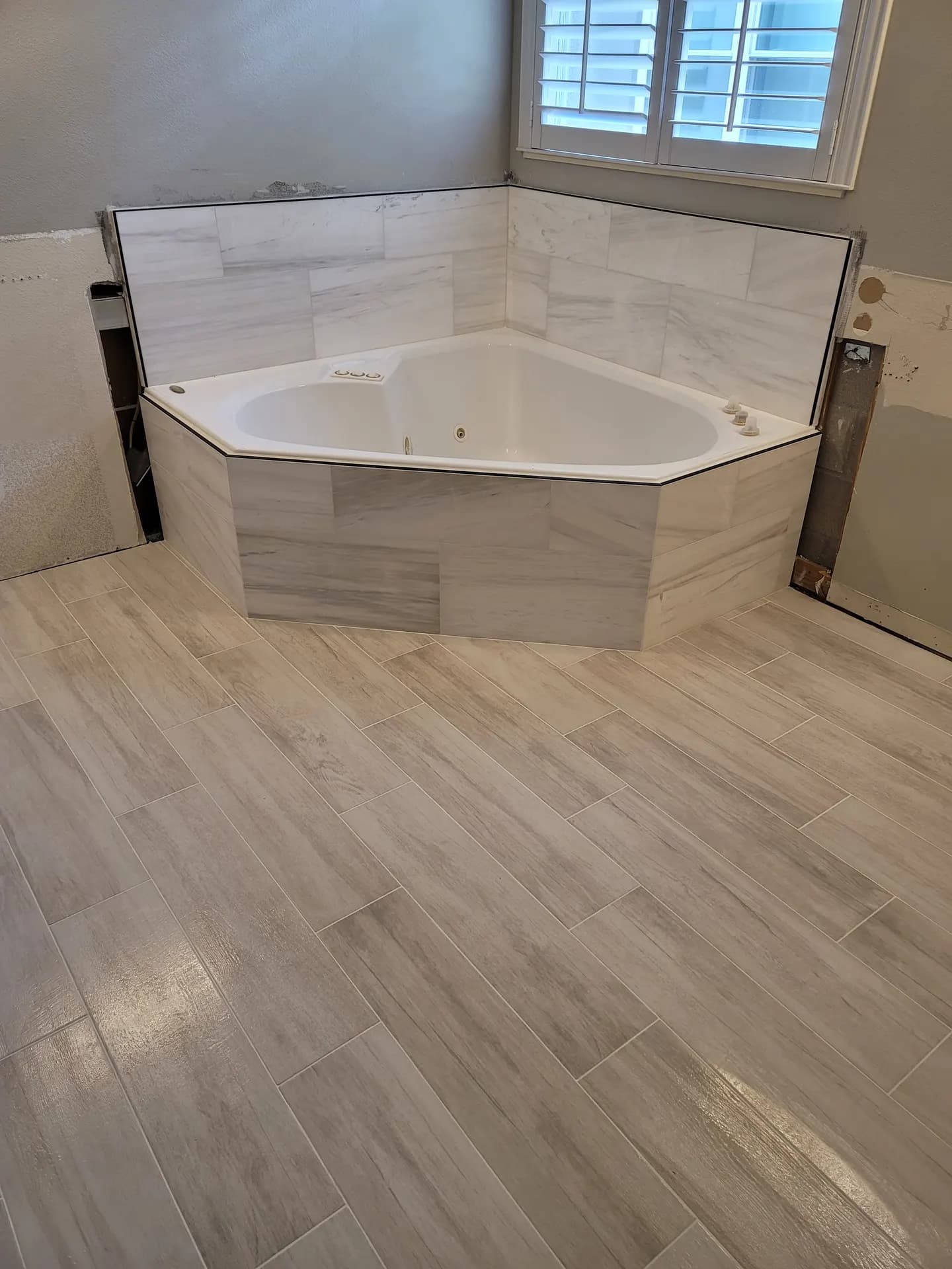 Corner soaking tub wrapped in marble-look tile with black metal trim, surrounded by wood-look porcelain plank flooring laid in an offset pattern.