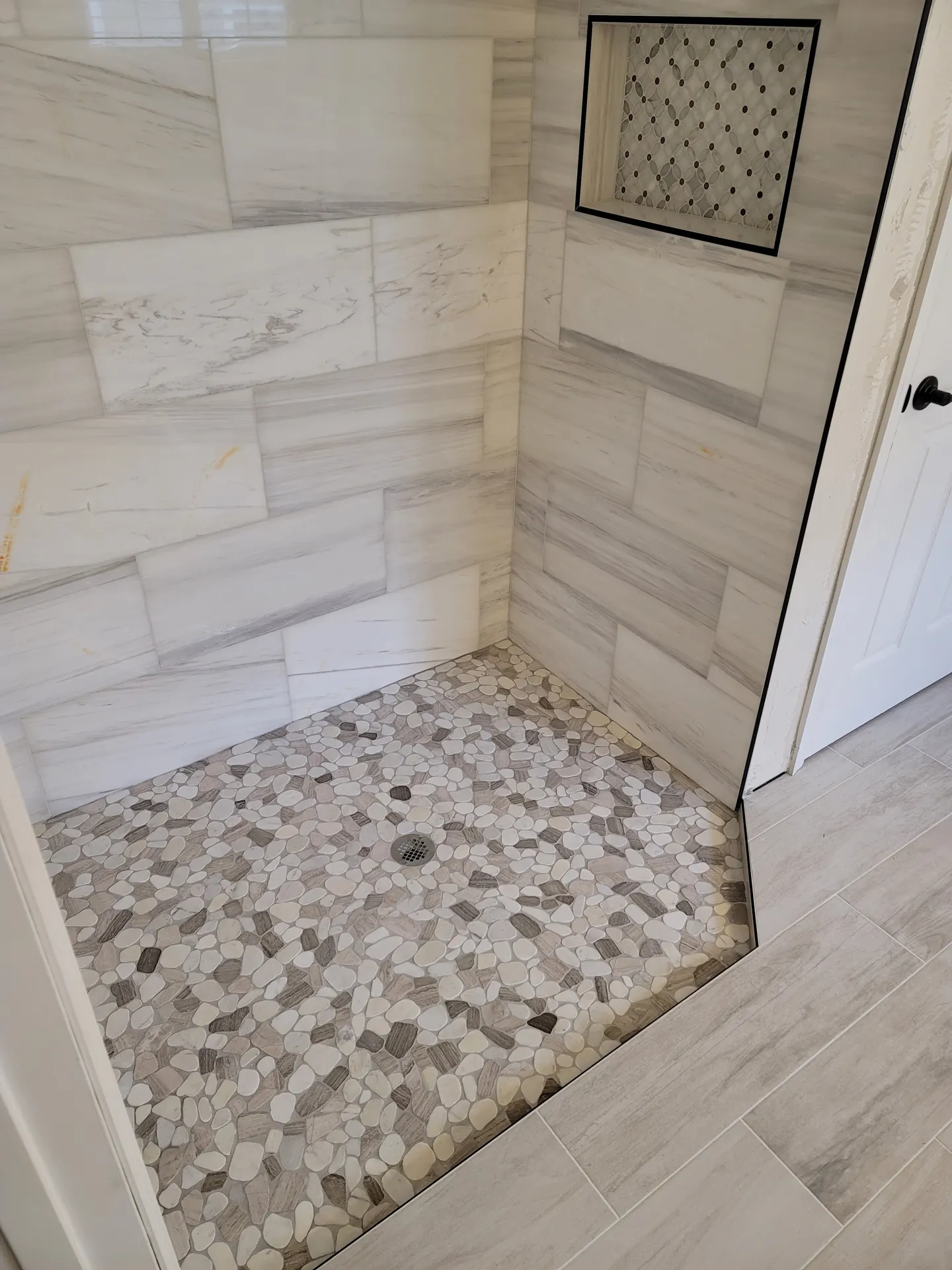 Walk-in shower with a taupe and cream pebble-stone floor, polished marble-look large-format wall tile, and a diamond mosaic inset niche.