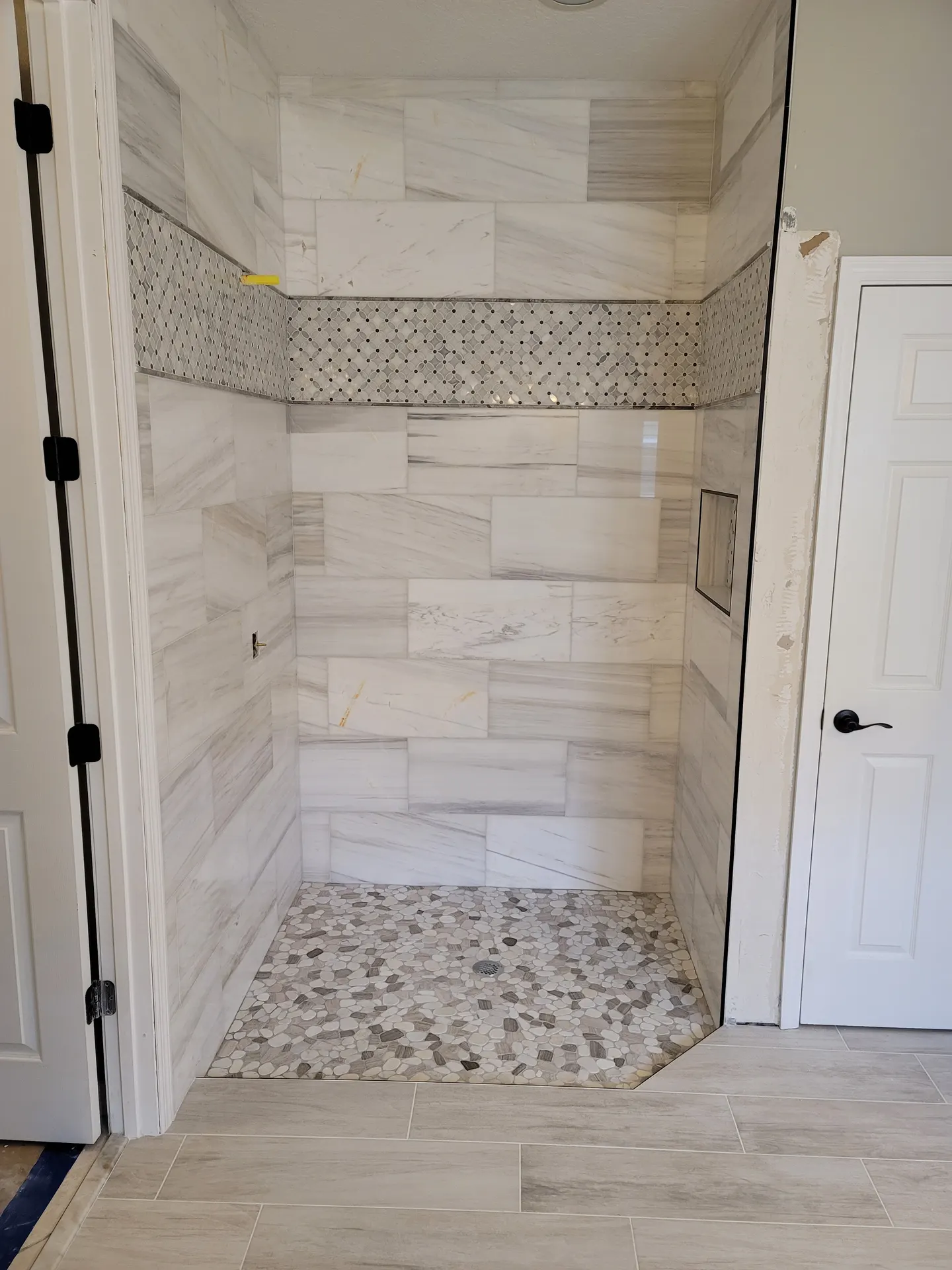 Master bathroom shower featuring straight-lay marble-look wall tile, a vertical floral mosaic accent band, and a multicolor pebble shower pan with drain.
