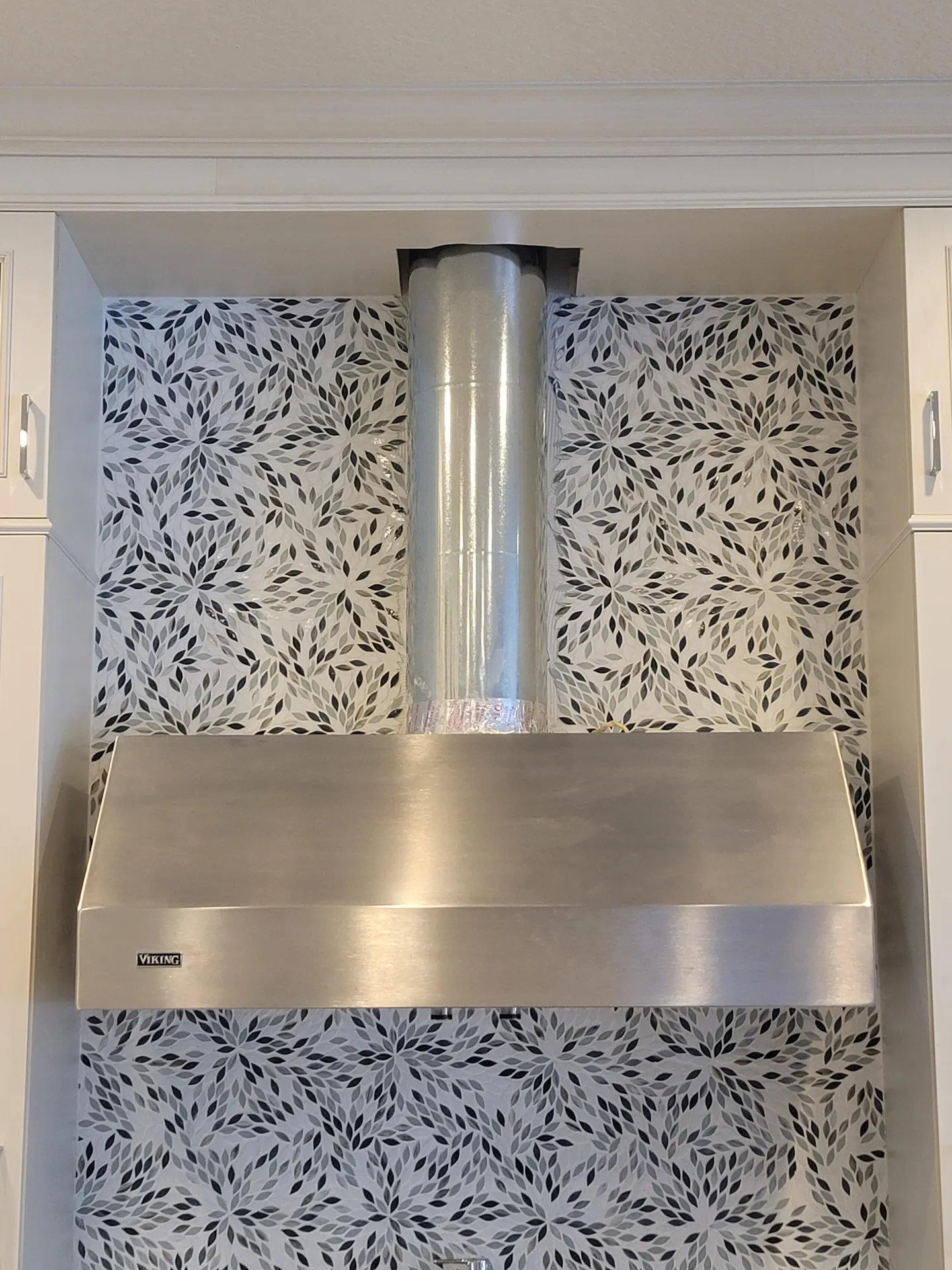 Kitchen range wall backsplash in a gray, black, and white flower-petal mosaic behind a stainless steel Viking hood with white cabinetry.