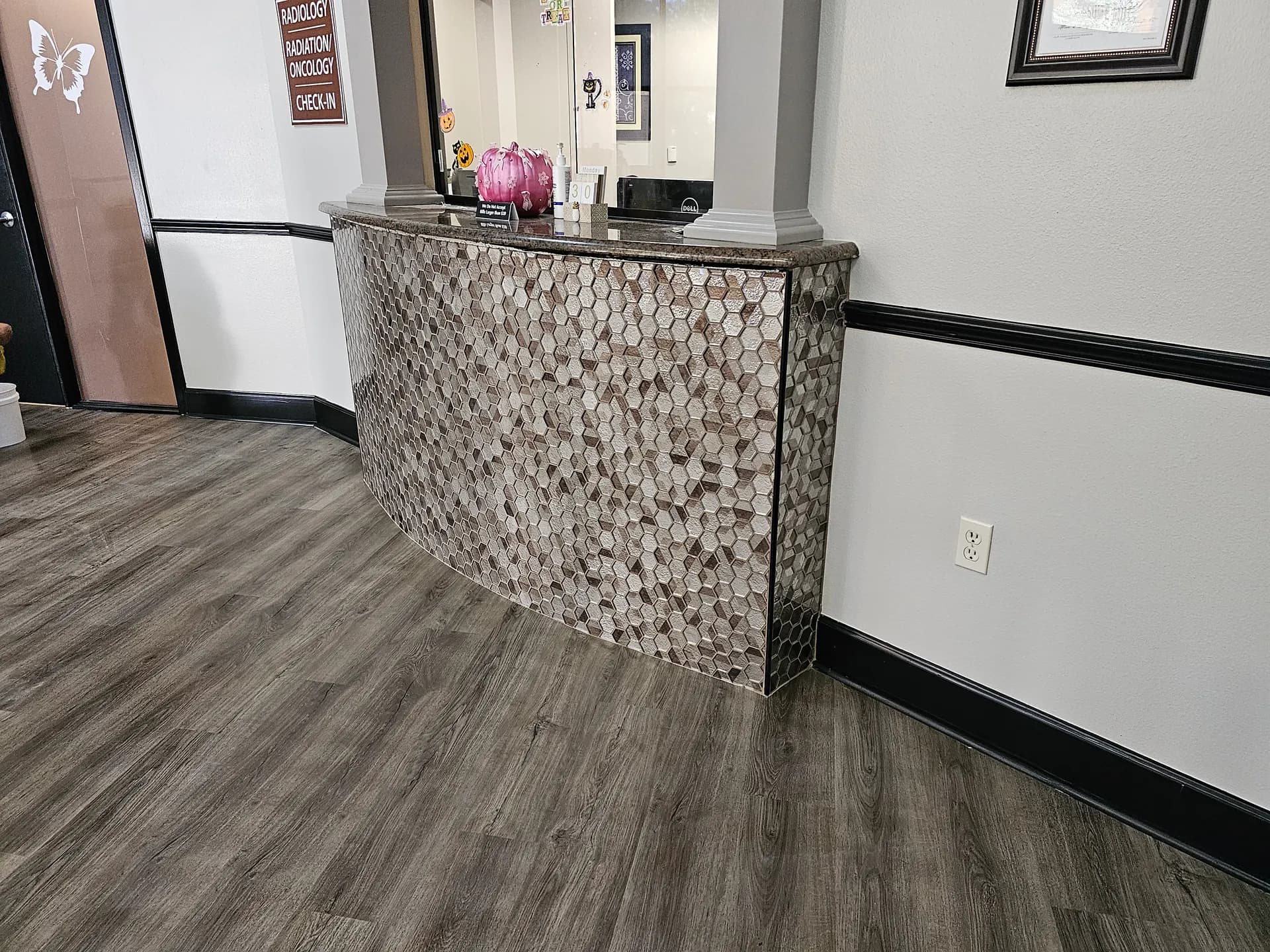 Angled side view of a curved reception desk clad in hexagon glass mosaic tile, highlighting the wrap-around edge and gray wood-look plank floor.