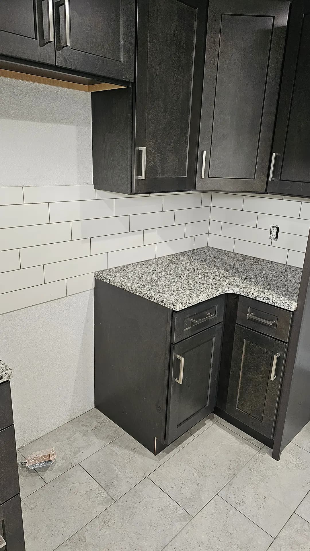 Large-format white subway tile backsplash laid in a vertical offset pattern behind a speckled granite countertop, dark gray shaker cabinets, and gray tile flooring in a kitchen under renovation.