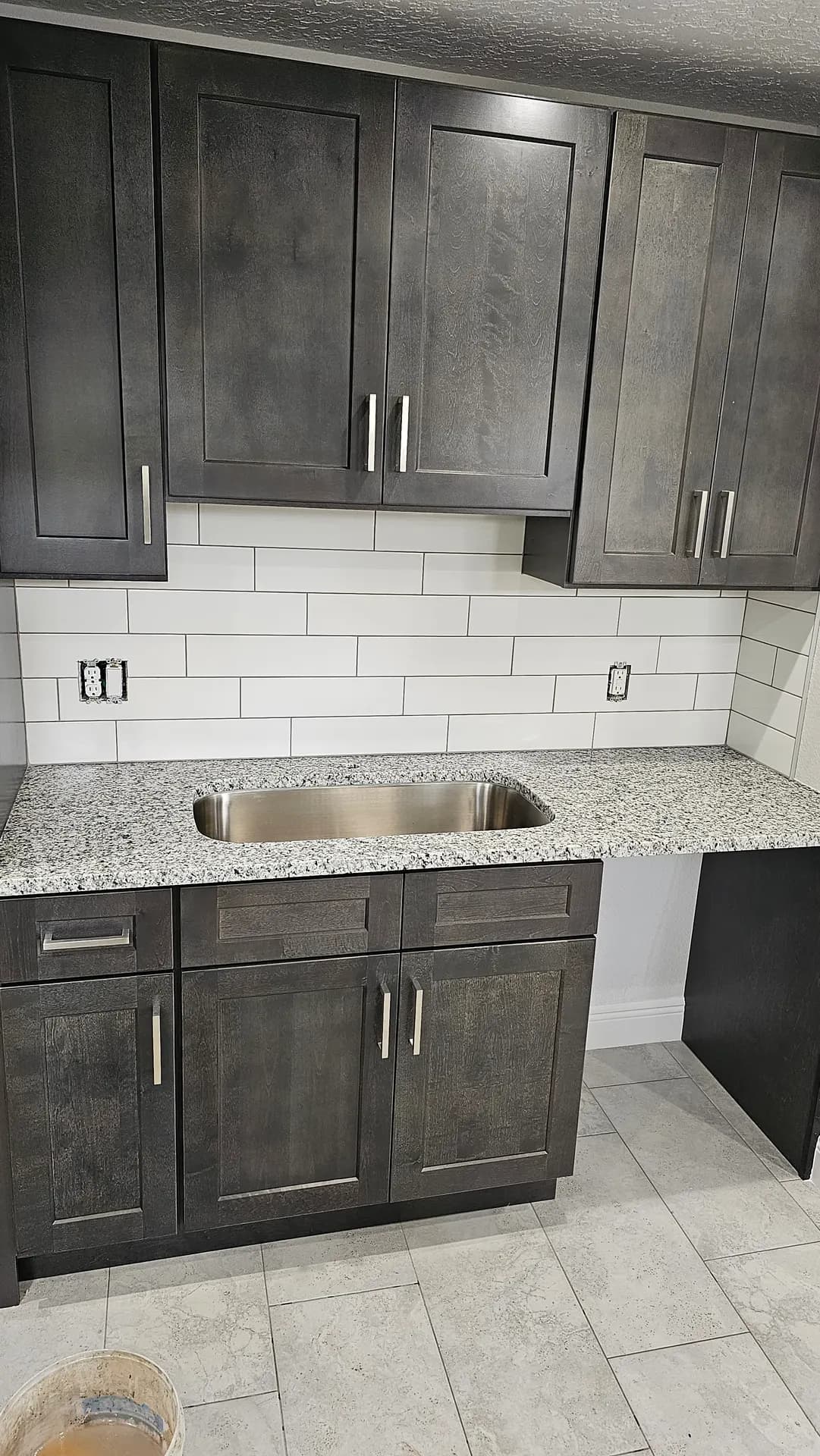 White subway tile installed vertically on a kitchen wall between dark gray shaker cabinets and a granite backsplash panel with brushed metal trim.