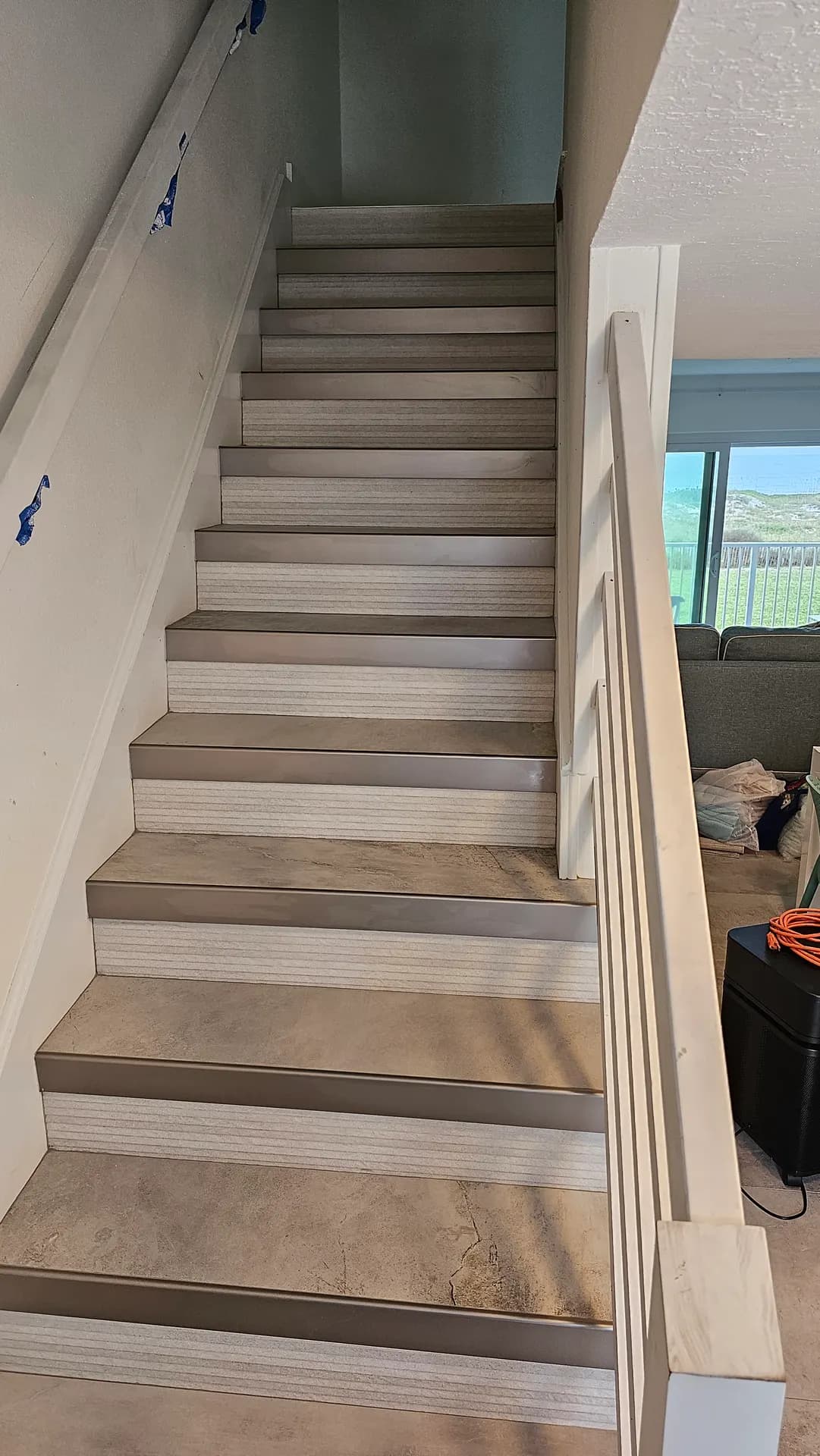 Full view of a staircase clad in large-format porcelain tile with metal stair nosing on every step, descending from a living area with a white wood railing.