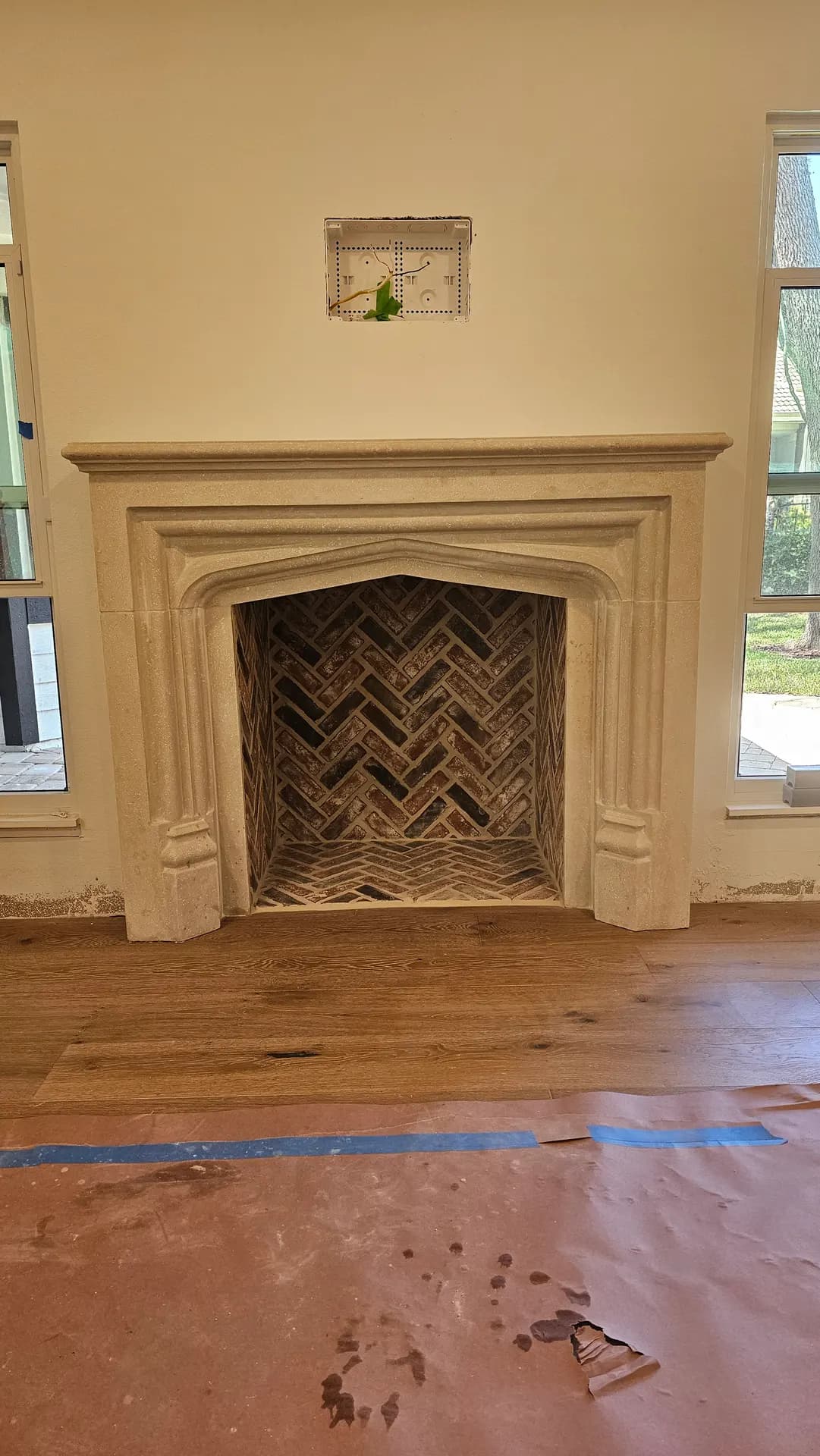 Cast stone fireplace surround with a herringbone reclaimed-brick firebox interior beside wide-plank wood flooring in a living room mid-remodel.