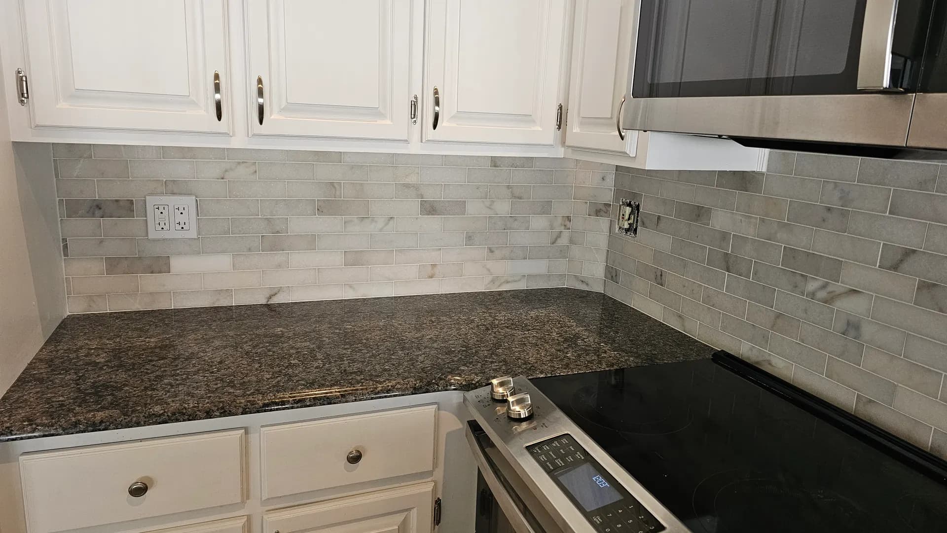 Marble subway tile backsplash wrapping a kitchen corner above dark granite counters with white raised-panel cabinetry and a stainless microwave hood.