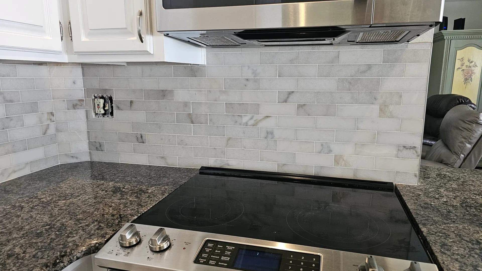 Marble subway tile backsplash behind a stainless steel cooktop and vent hood with dark granite countertops in a finished kitchen.