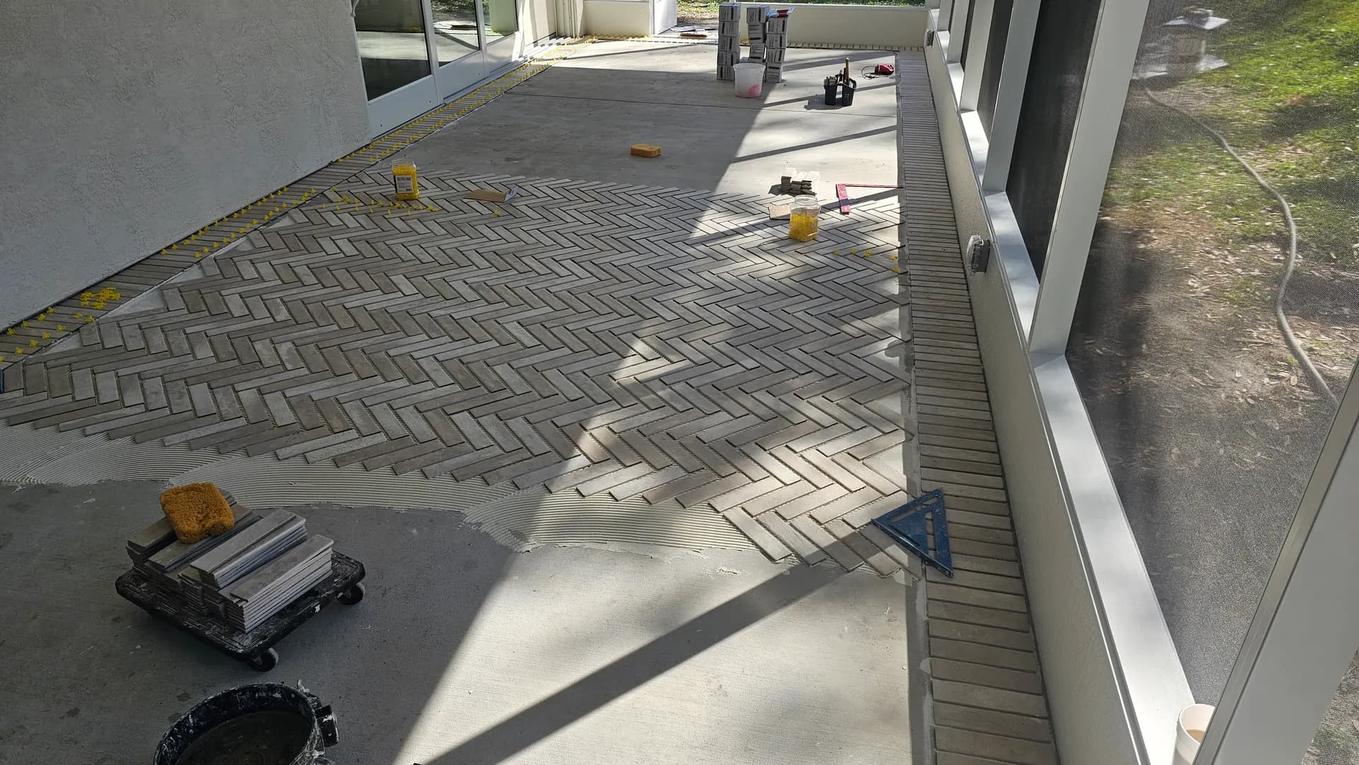 Brick-look porcelain tile being installed in a herringbone pattern with a straight-lay border on a screened lanai patio floor.